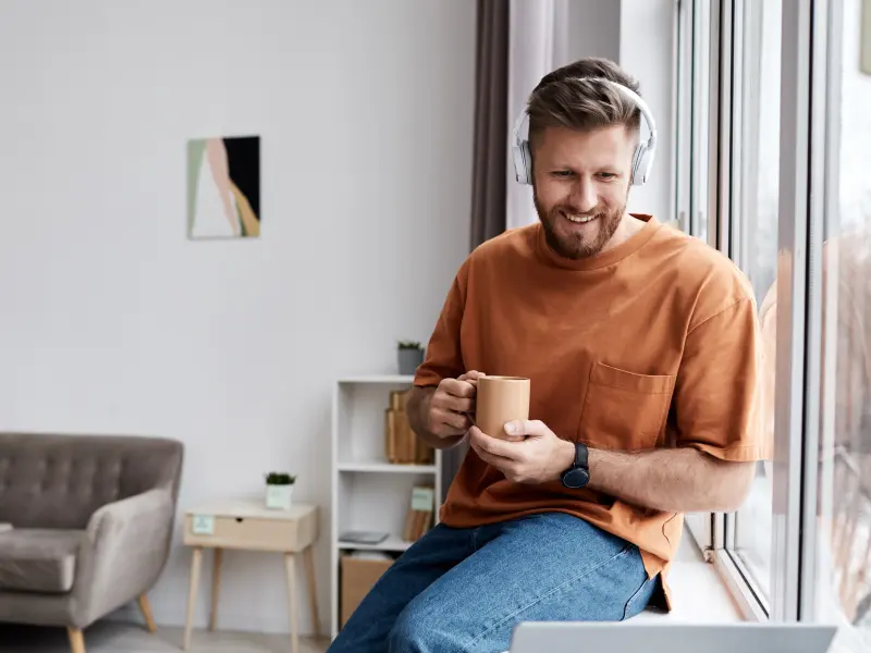 Ein glücklicher junger Mann mit einer Tasse Kaffee sitzt am Fenster Ein glücklicher junger Mann mit einer Tasse Kaffee sitzt am Fenster