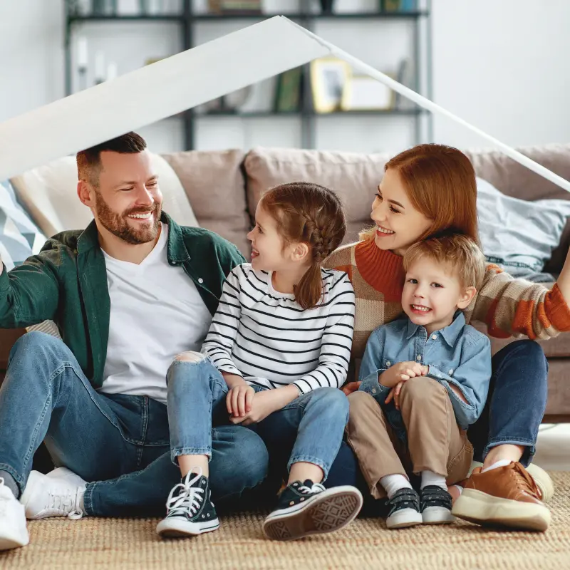 Familie sitzt vor ihrem Sofa und hält sich ein gebasteltes Dach über den Kopf Familie sitzt vor ihrem Sofa und hält sich ein gebasteltes Dach über den Kopf