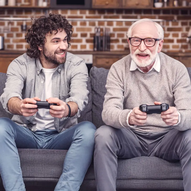 BarmeniaGothaer Risikolebensversicherung: Ein junger Mann sitzt mit einem älteren Mann auf einem Sofa und spielt auf einer Gaming-Konsole BarmeniaGothaer Risikolebensversicherung: Ein junger Mann sitzt mit einem älteren Mann auf einem Sofa und spielt auf einer Gaming-Konsole