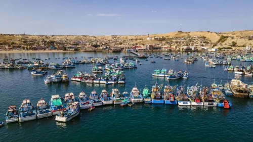 Aerial view of a crowded harbor with many colorful fishing boats docked and floating on the water, with a coastal town and arid hills in the background under a clear blue sky.