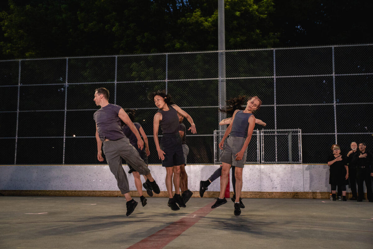 three dancers jumping while looking away from each other. They are wearing different shades of grey. There are three more dancers behind them whose faces cannot be seen. Behind them is a fence and green trees.