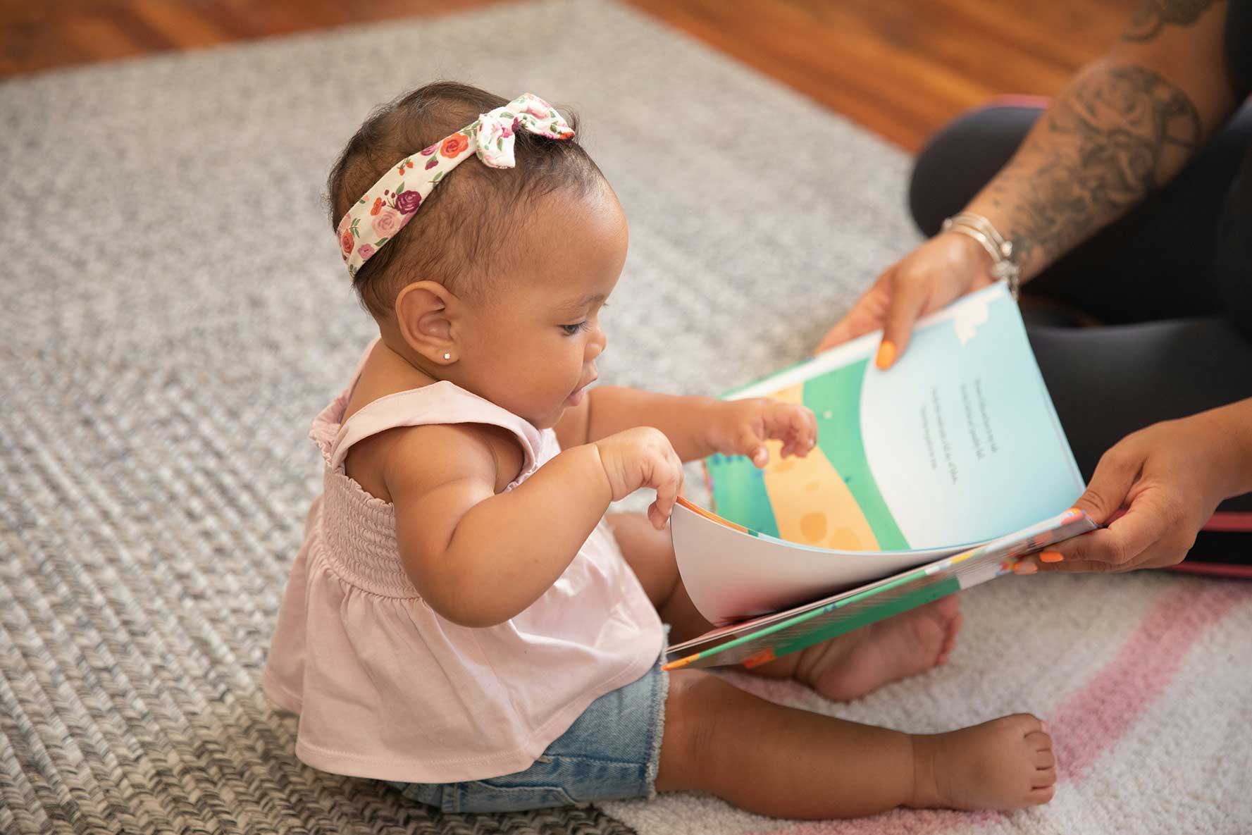 Baby sitting on floor turning the pages of a book with the help of the arms of a caregiver.