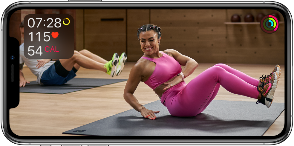 Una pantalla mostrando un entrenador que imparte un entrenamiento de Apple Fitness Plus. En la esquina superior izquierda se muestra información sobre la duración del entrenamiento, la frecuencia cardiaca y las calorías quemadas. En la esquina superior derecha se muestra el progreso de los círculos de los objetivos de Moverse, Ejercicio y Pararse.