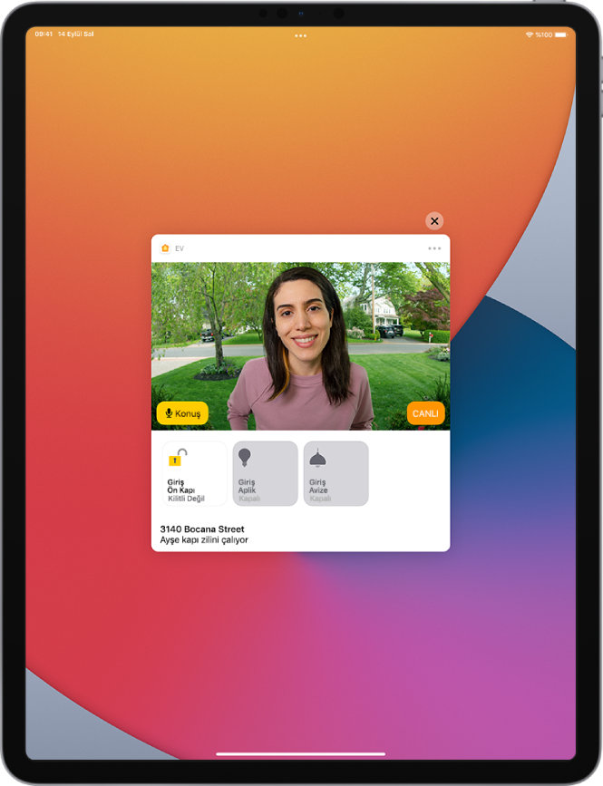 iPad ekranında Ev uygulamasından bir bildirim var. Sol tarafta bir Konuş düğmesiyle ön kapıdaki bir kişinin resmi gösteriliyor. Alt tarafta ön kapı ve giriş ışıkları için aksesuar düğmeleri var. En altta “Ayşe kapı zilini çalıyor” sözcükleri ve ev adresi var. Bildirimin sağ üst tarafında Kapat düğmesi var.