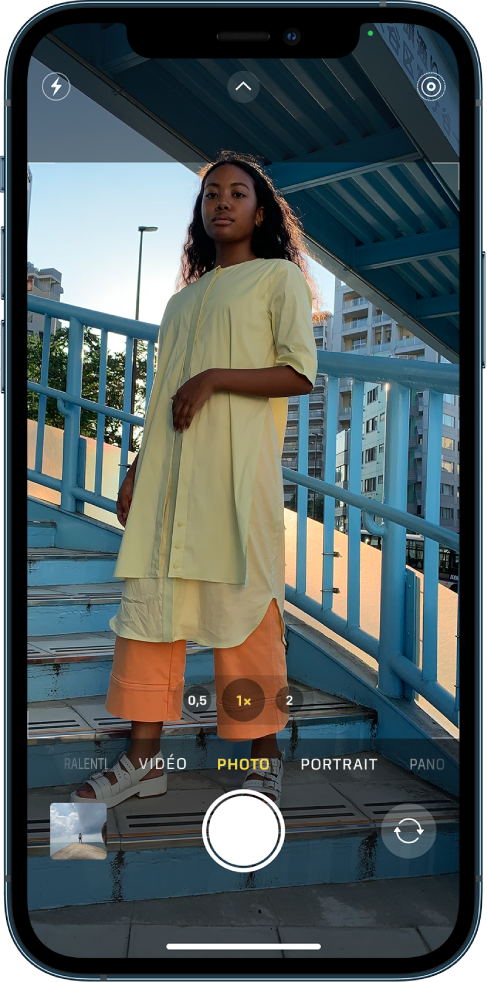 Appareil photo en mode Photo, avec les autres modes à gauche et à droite sous le viseur. Les boutons pour le flash, les commandes de l’appareil photo et le mode Live Photo apparaissent en haut de l’écran. Le bouton du visualiseur de photos et de vidéos se trouve dans le coin inférieur gauche. Le bouton « Prendre une photo » se trouve en bas au centre et le bouton « Sélecteur de caméra - face arrière » est dans le coin inférieur droit.