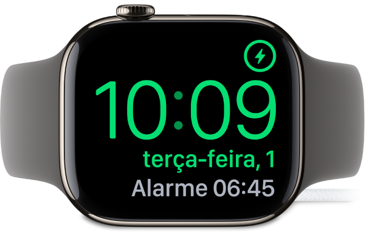 Um Apple Watch colocado de lado e ligado ao carregador, com o ecrã, com o símbolo de carregamento no canto superior direito e, por baixo, a hora atual e a hora do próximo alarme.