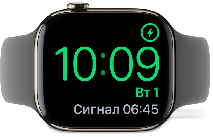 Apple Watch покладено на бік і підʼєднано до зарядного пристрою. На екрані відображається значок заряджання у верхньому правому куті, під ним — поточний час і час наступного будильника.