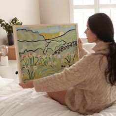 a woman sitting on top of a bed holding up a painting in front of her