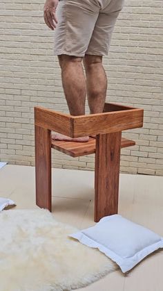 a man standing on top of a wooden table next to a white rug and pillow