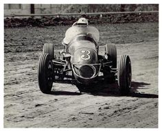 an old photo of a man driving a race car down a dirt road in front of a building