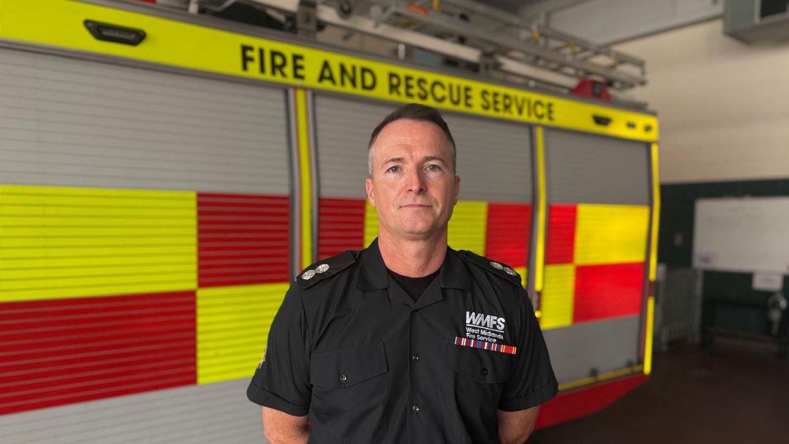 A man in a black shirt with 'West Midlands Fire Service' on it looks at the camera. He is standing in front of a fire engine in a garage.