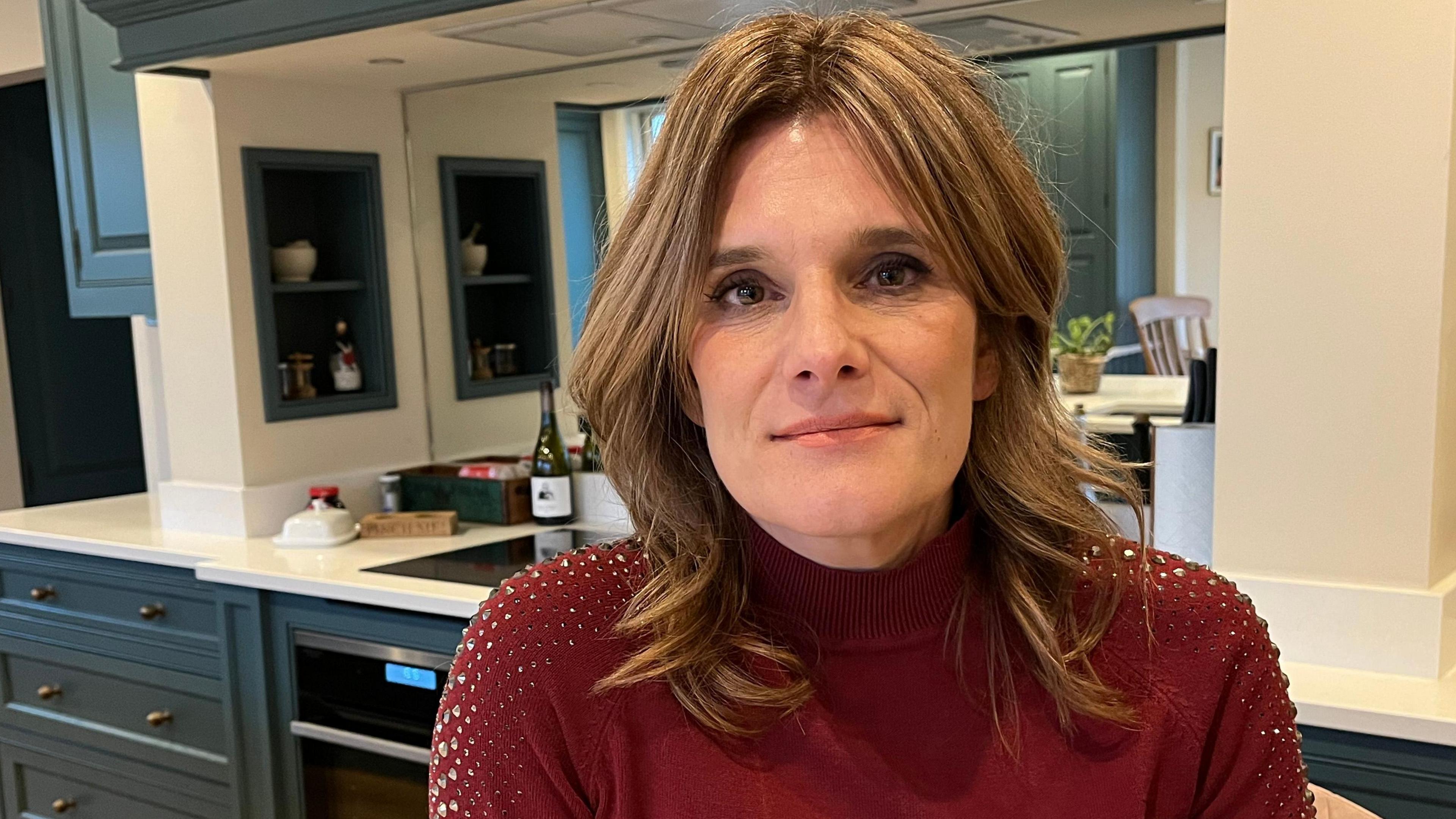 Ellen looks at the camera with a small smile. She is in a kitchen in a house, with the cabinets and work surface visible behind her. She has short dark blonde hair, which is loosely curled, and wears a red jumper with diamante sequins on the shoulders.