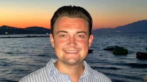 A white young man with brown hair, wearing a striped blue and white shirt. He's stood in front of a body of water with a sunset scene in the background.