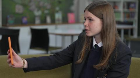 Imogen pictured sitting at her school, Appleton Academy in Bradford. She has long brown hair and is wearing a school blazer, jumper and white shirt. She is holding a green mobile phone out in front of her.
