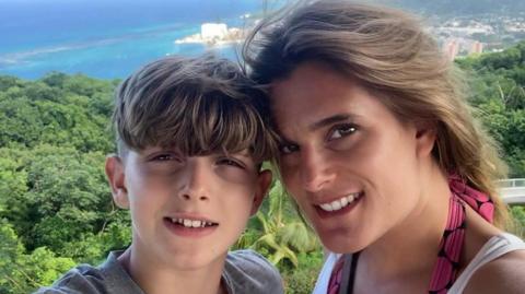 Jools Sweeney and his mother Ellen Roome. Both of them are looking ahead and smiling. There are trees and the sea behind them.