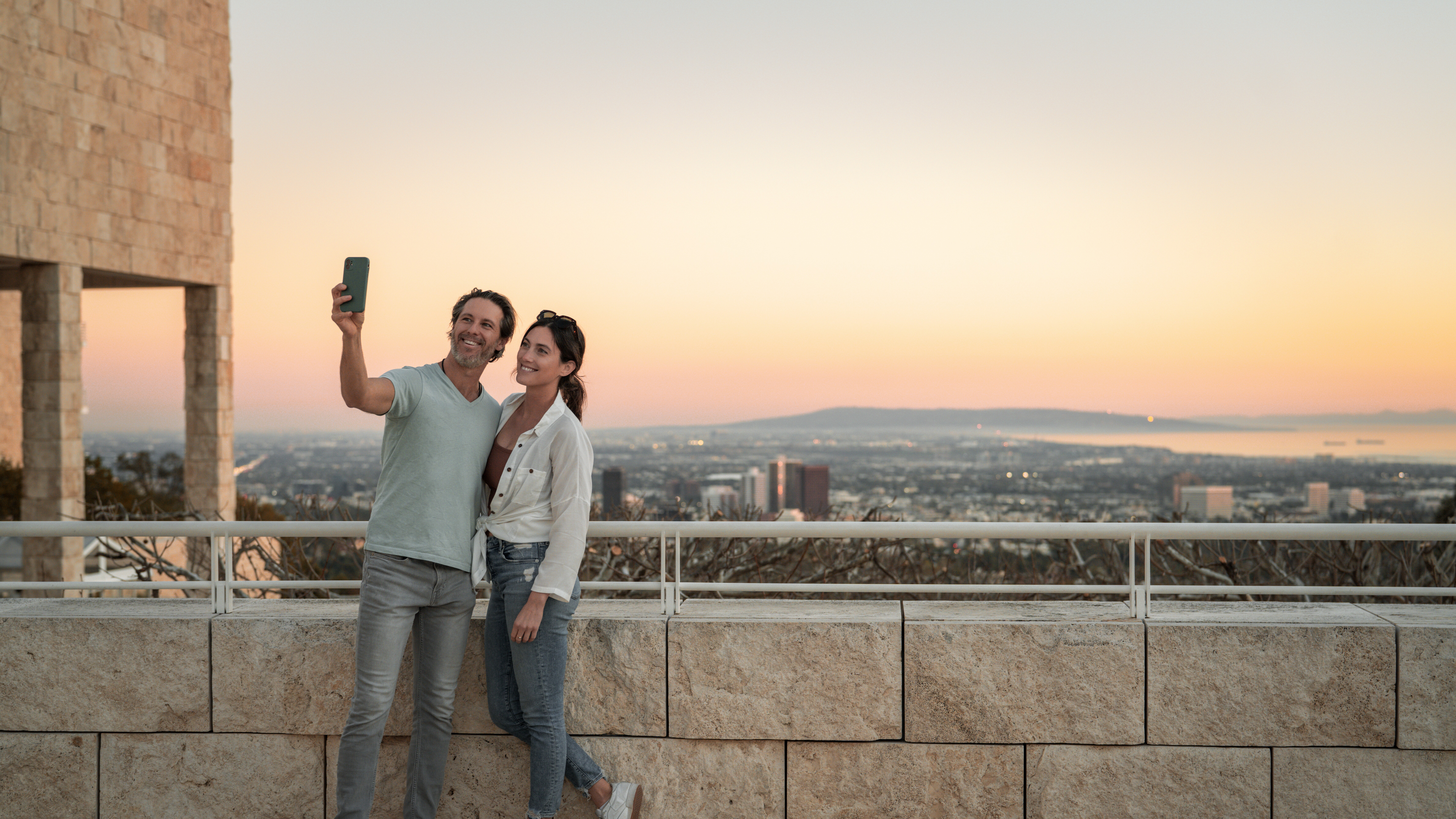 Two people pose for a selfie on a stone balcony overlooking a large city view during sunset