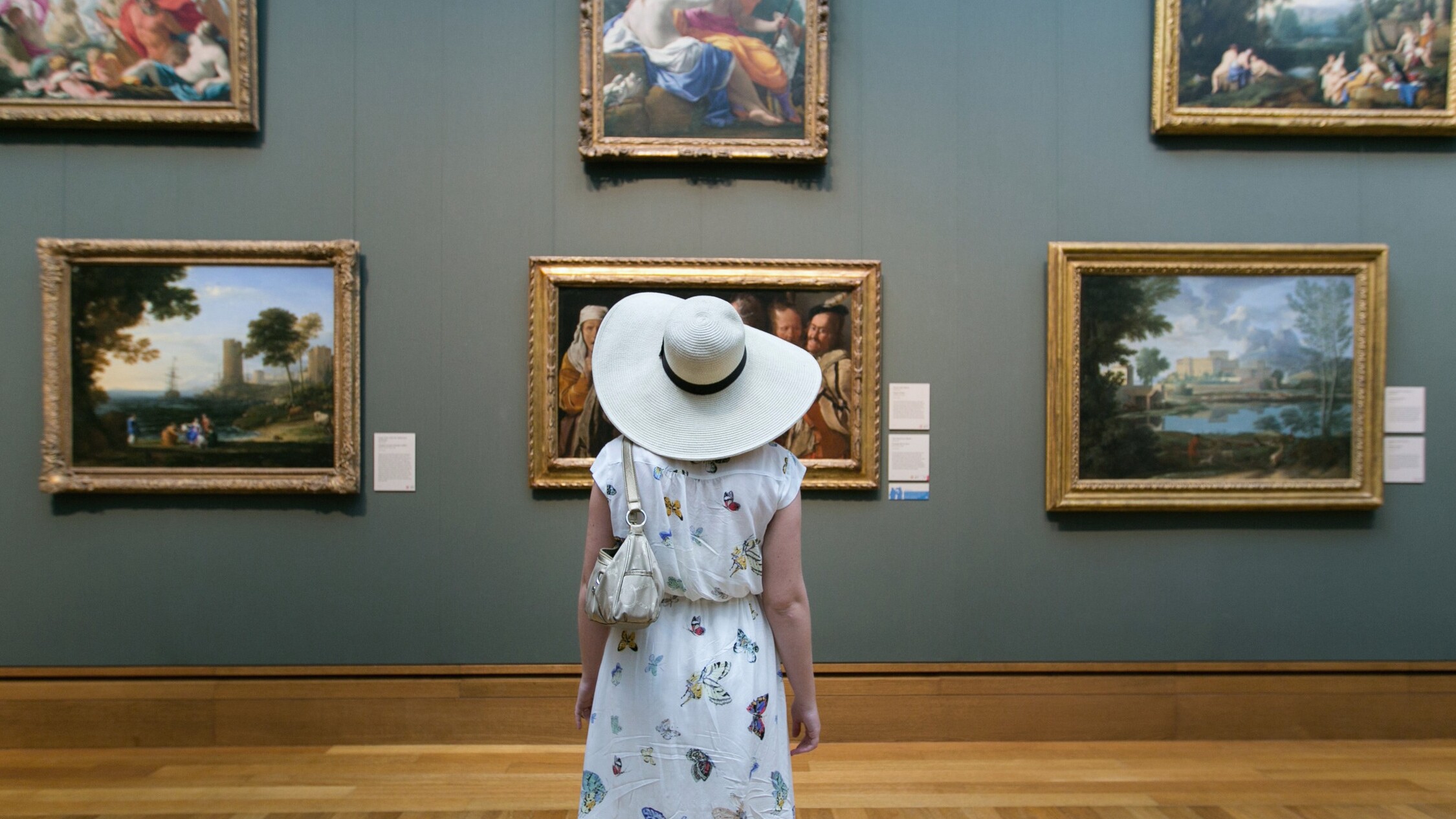 Wearing a dress covered in butterflies and a wide-brimmed white hat, a visitor looks at a wall of paintings.