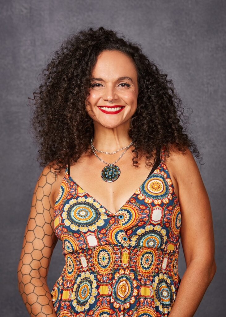 Rebeca Marrero. Woman with dark brown curly hair, light brown skin, smiling, wearing a colorful patterned dress.