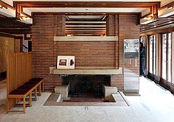 The fireplace at the eastern end of the living room. The mantelpiece is made of stone, and the fireplace is flanked by brick columns. There is a gap between the columns above the fireplace, and the dining room is visible through the gap.