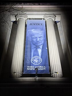Nighttime photograph of a large vertical banner hanging between two neoclassical limestone columns. The banner primarily shows Donald Trump's face, with "U.S. Department of Justice Robert F. Kennedy Building" above and "Make America Safe Again" below.