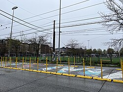 A mural is on the street surrounded by yellow barricades with soccer fields in the background. The mural's M has a white background, covered in splashes of blue, teal, yellow, and purple paint. Part of an E,S and A are shown: the S is colored in red, black, and white.