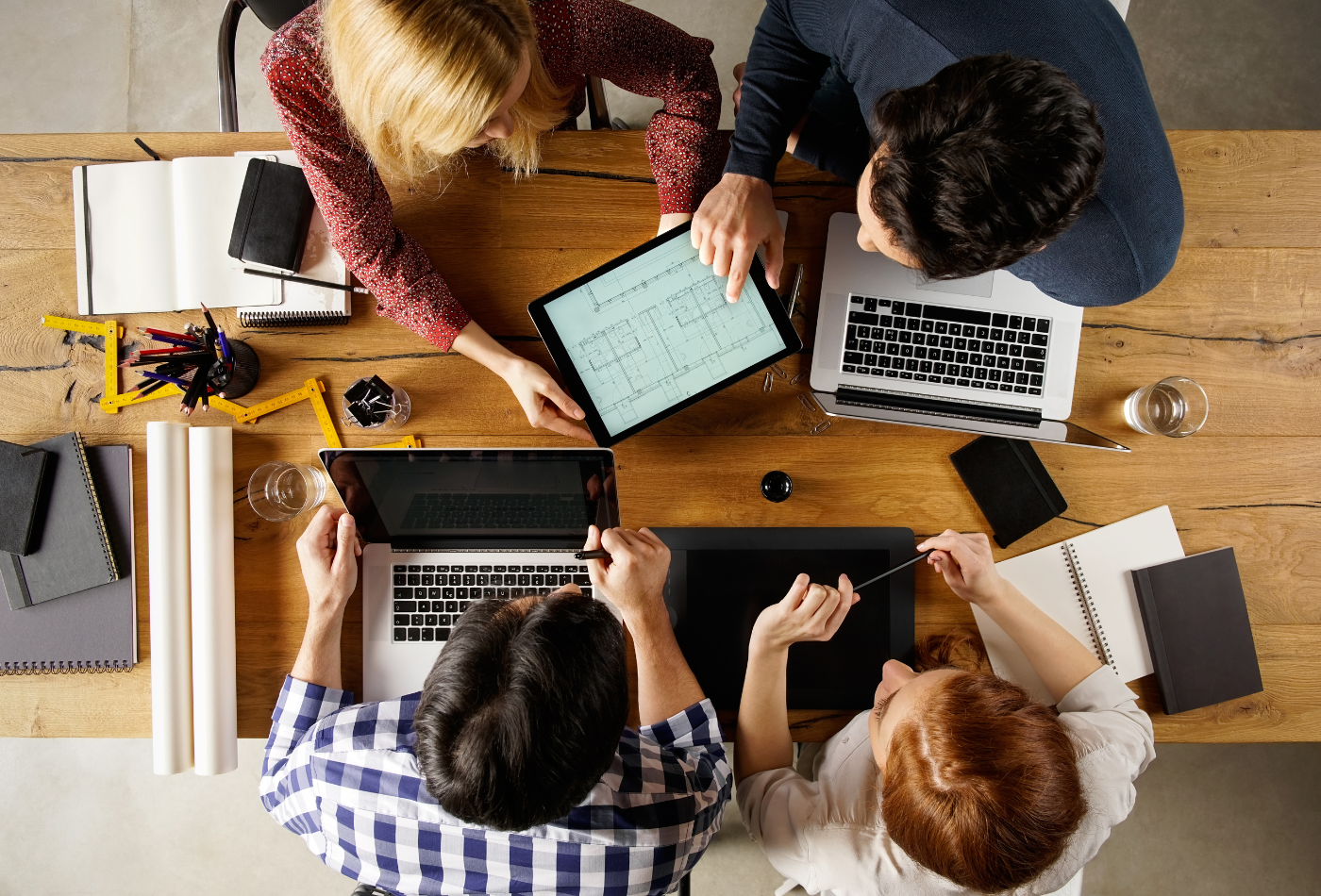 Five people sitting around a wooden table, collaborating. They are using laptops, tablets, and notebooks. There are architectural blueprints on a tablet and several stationery items scattered on the table.