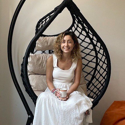 Eine lächelnde Frau mit lockigem Haar sitzt in einem schwarzen Hängesessel und hält eine Tasse, neben ihr ein oranger Sitzsack.