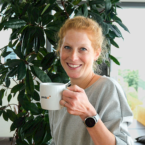 Alisha Westbomke, Recruiting Specialist bei jambit, lächelt in die Kamera und hält eine Kaffeetasse mit jambit-Logo in einem modernen Büro.
